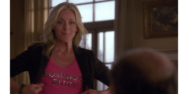 Jenna Maroney from 30 Rock wearing a bright pink shirt with rhinestone lettering and a black jacket, striking a confident pose in an elegant room.