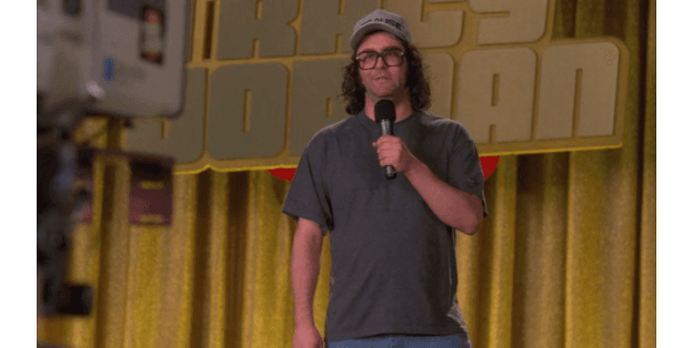 Frank Rossitano from 30 Rock performing standup comedy, wearing his signature hat and glasses with a casual gray t-shirt.