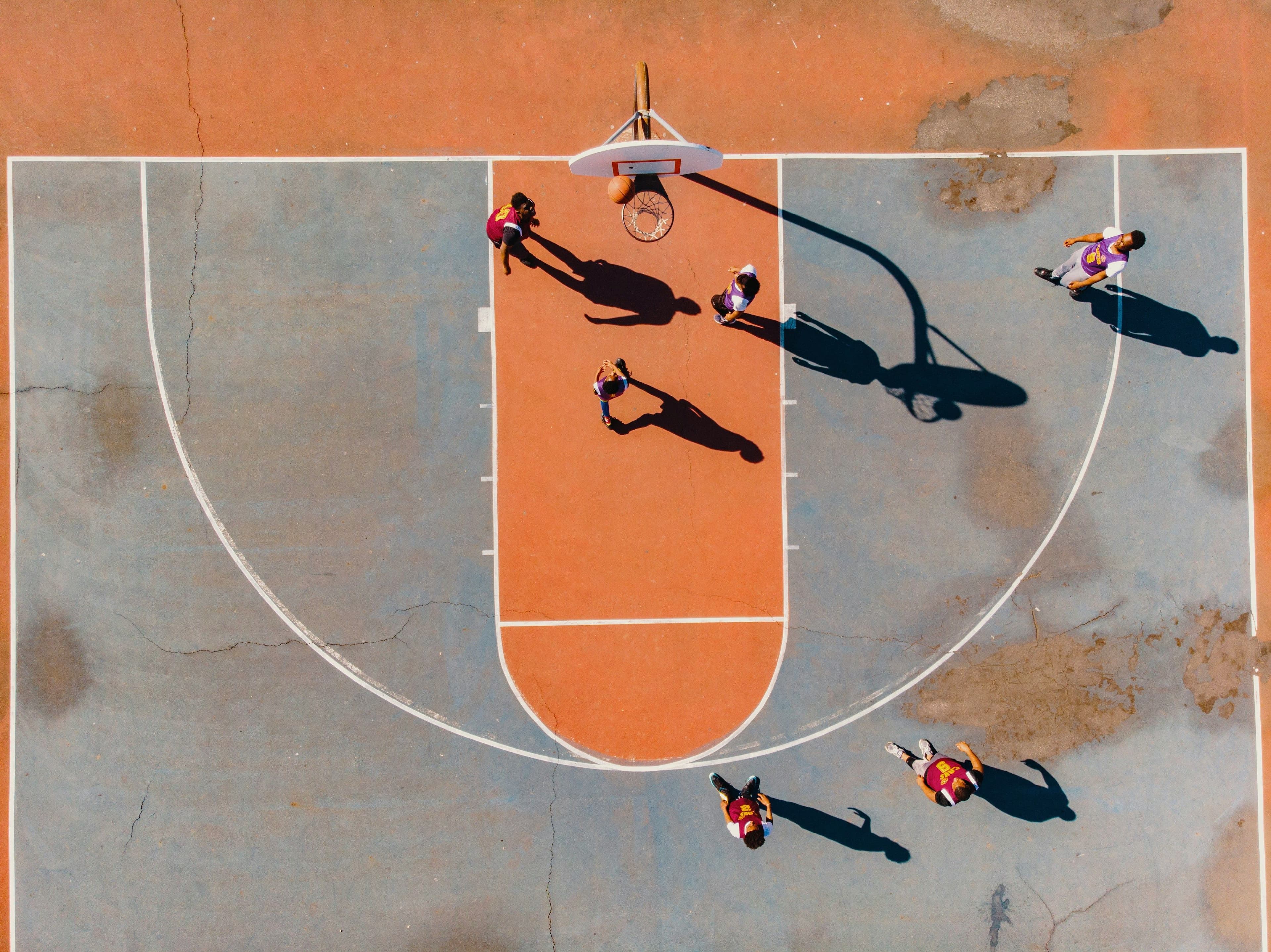 Aerial drone view of a basketball court with orange and gray surface showing players in red and purple jerseys during a pickup game. The image captures long shadows cast by players and the basketball hoop, highlighting the outdoor recreational nature of March Madness season.