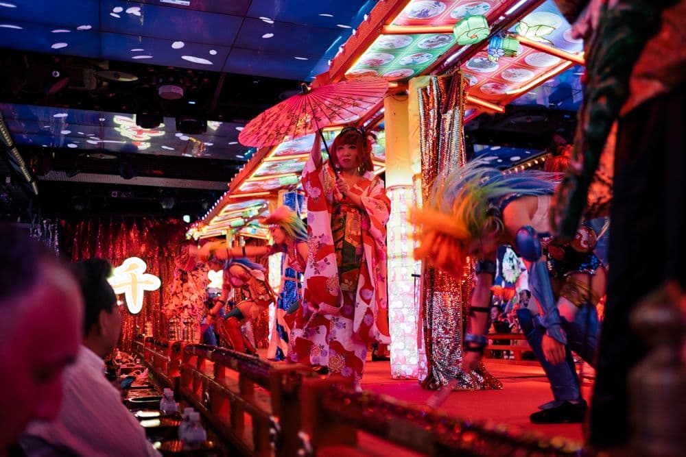 A performer in a traditional Japanese kimono holding a red umbrella stands on an elaborately decorated, brightly lit stage at the Robot Restaurant. The performer is framed by colorful illuminated panels and strings of beads, while audience members can be seen sitting below.