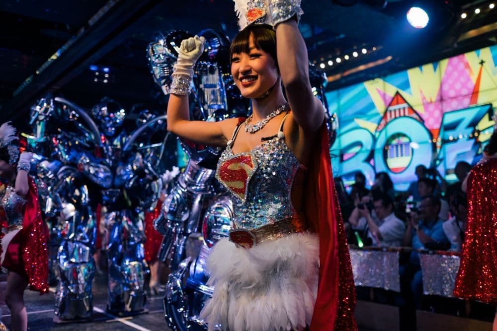 A performer in a sparkly superhero-inspired costume with a red cape and crystal embellishments smiles joyfully on stage at the Robot Restaurant in Tokyo. She's raising her arms in celebration with robotic figures visible in the background and colorful digital screens displaying vibrant graphics.
