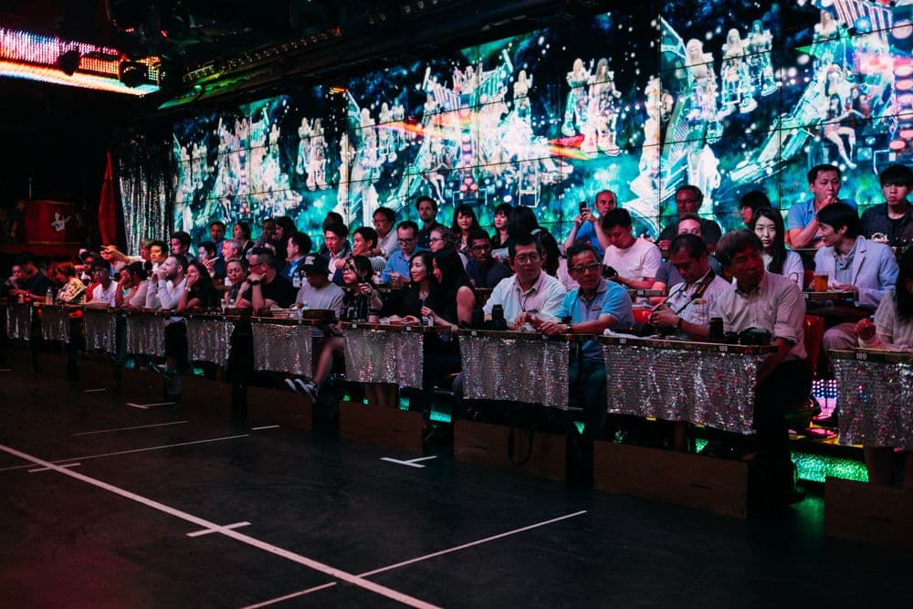 The audience at Tokyo's Robot Restaurant sitting along sparkly barricades with tables in front of them. Behind them is a large digital wall display showing illuminated figures. The crowd appears engaged and many are taking photos of the performance.