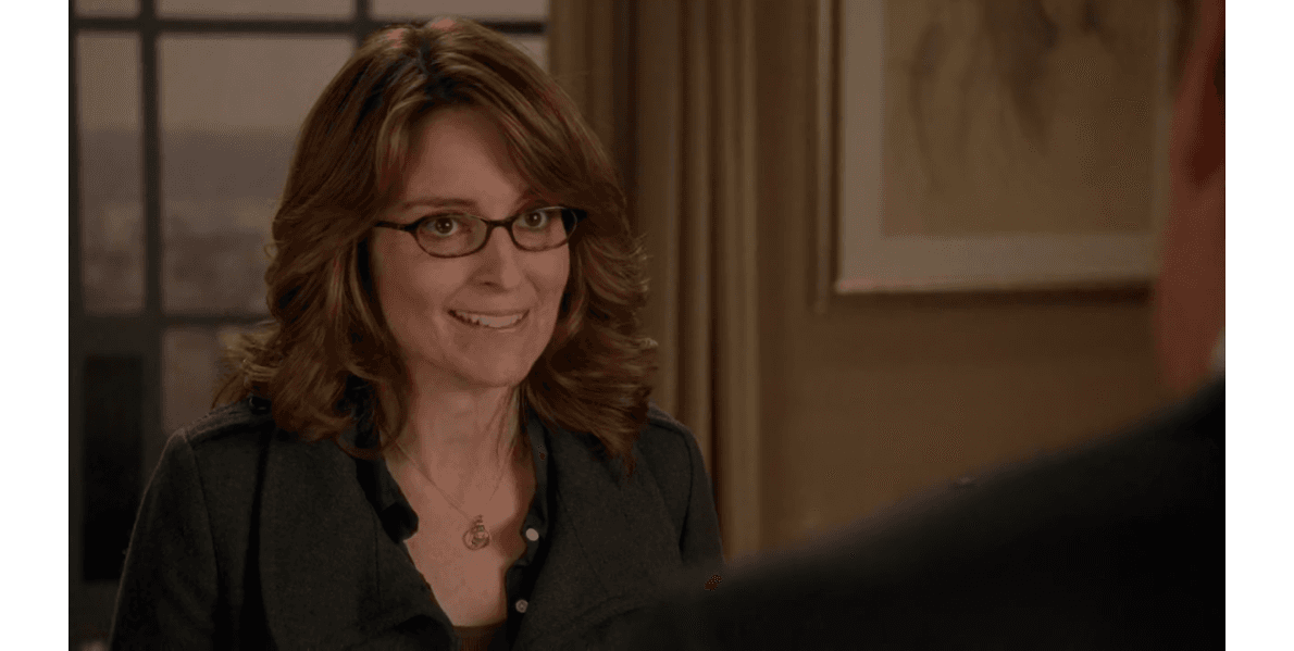 Liz Lemon from 30 Rock smiling during a conversation, wearing her trademark glasses and a dark cardigan over a button-up shirt with a subtle necklace, in an office setting with warm lighting.
