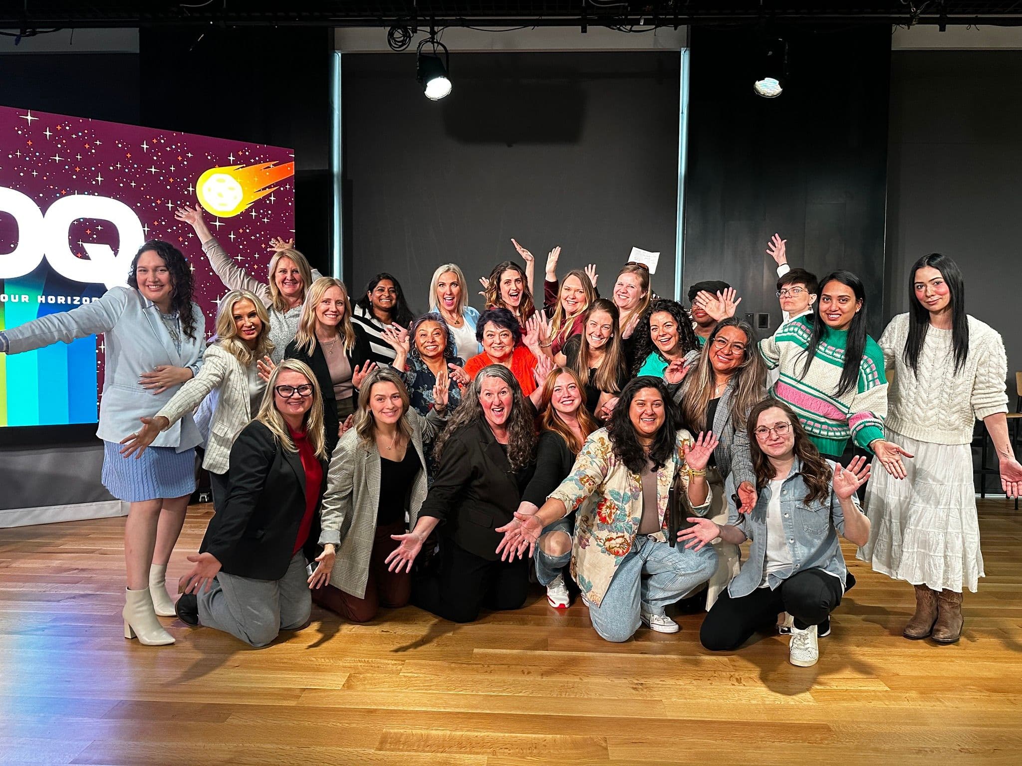 A large group of diverse women celebrating at a Tech-Moms program graduation event, posing enthusiastically together on stage with hands raised in celebration beneath a colorful 'Expand Your Horizons' banner.