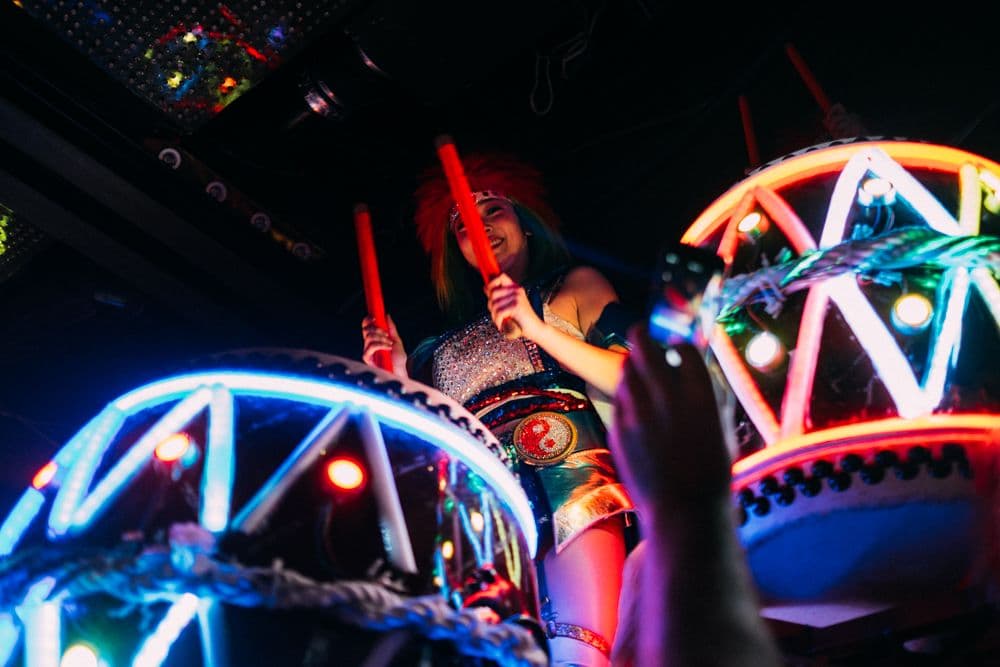 A performer with colorful hair wearing a sparkly costume holds glowing red drumsticks above geometric neon structures at Tokyo's Robot Restaurant. The image captures the vibrant, high-energy atmosphere with bright LED-lit stage elements.
