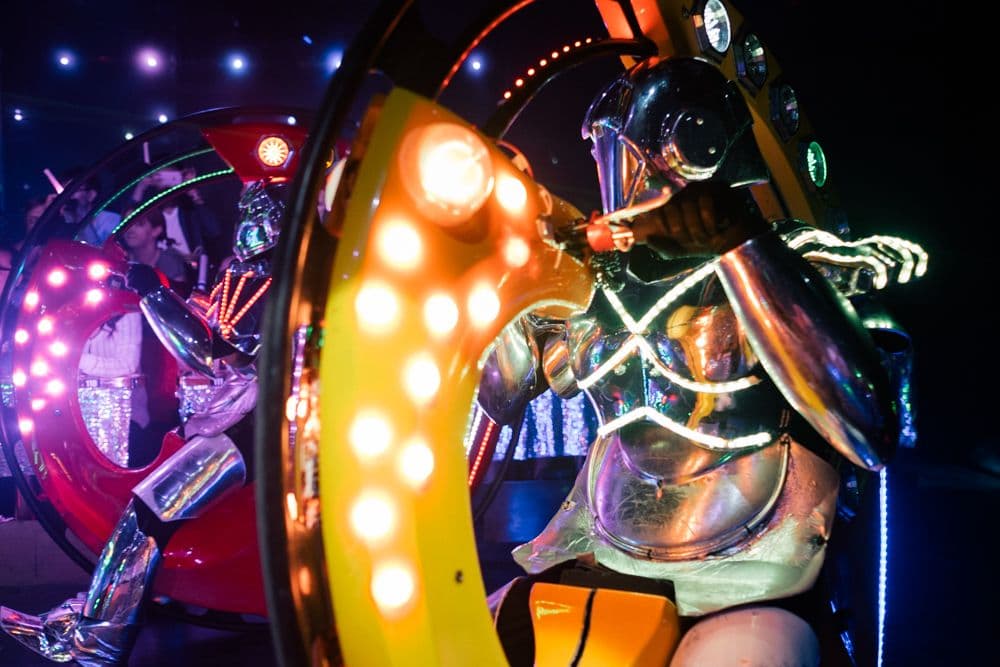 A futuristic robot performer in metallic armor is illuminated by green and orange lights at Tokyo's Robot Restaurant. The robot is mounted on a colorful platform with LED lights while excited audience members can be seen in the background behind a sparkly barricade.